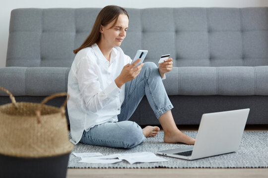 Image of attractive concentrated woman wearing white shirt and jeans sitting on floor near sofa, using laptop and phone, holding credit card, checking data for online paying.
