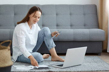 Portrait of Caucasian woman wearing white shirt and jeans sitting on floor near sofa, using laptop,...