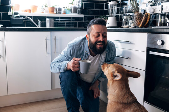 Happy man with coffee cup looking at dog in kitchen