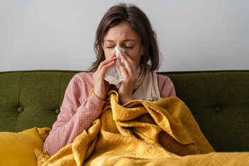 Sick woman blowing nose in facial tissue sitting on sofa at home