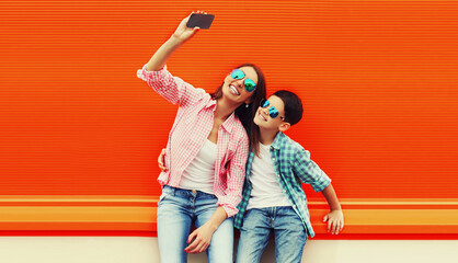 Happy smiling mother with son teenager taking selfie by smartphone on colorful orange background
