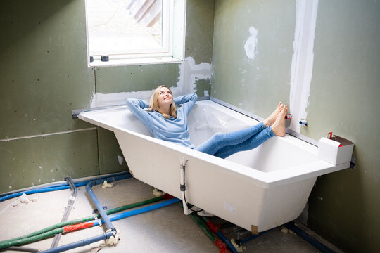 Happy blond woman with hands behind head sitting in bathtub in attic