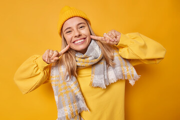 Glad young woman points at toothy smile has white even teeth wears hat sweatshirt and scarf around neck feels very happy isolated over yellow studio background. Human facial expressions concept