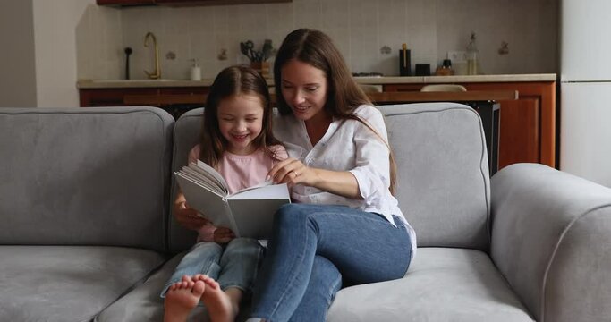 Loving mom read book her little daughter seated on sofa at home. Preschool girl enjoy pastime and fascinating fairytale rest on couch with caring mother. Kids development, intellectual growth concept