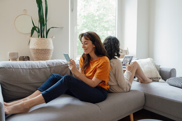 Smiling woman using smart phone sitting back to back with daughter using tablet PC on sofa at home
