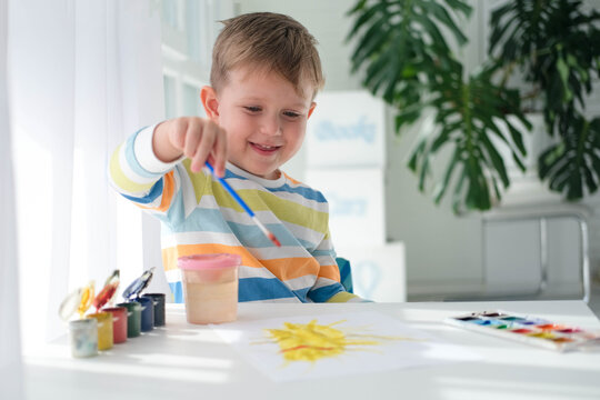Happy kid painting with brush and colorful paints. A little boy draws a picture with paints on a sheet of paper. The child is learning to draw. Home creativity.