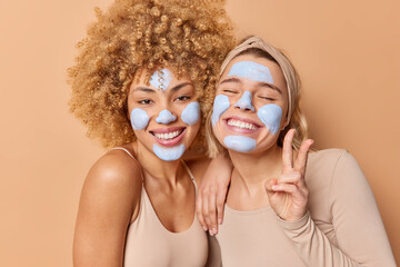 Positive carefree young women with beauty nourishing mask on face stand closely to each other smile...