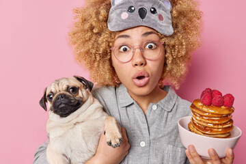 Scared young curly haired woman poses with pancakes with raspberries and pug dog going to have breakfast dressed in pajama and sleepmask cannot believe her eyes isolated over pink background.