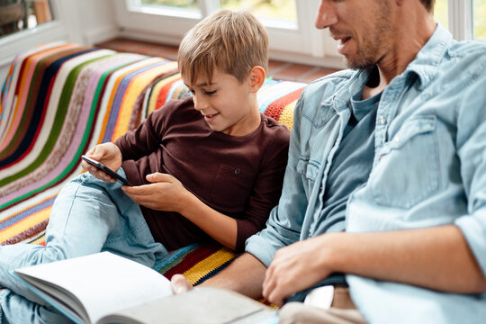 Man with book looking at son using smart phone sitting on sofa ta home