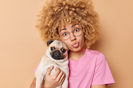 Lovely young woman poses with pug dog expresses love and care to favorite pet dressed in casual t shirt spends free time with new family member isolated over brown background. Domestic animals