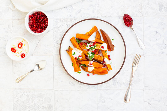 Plate of sweet potatoes with parsley, pomegranate seeds and cumin yogurt lying on white tiles