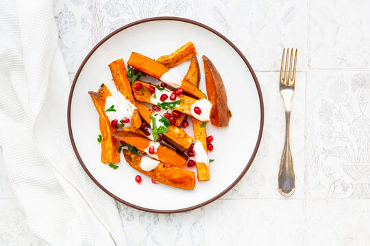Plate of sweet potatoes with parsley, pomegranate seeds and cumin yogurt lying on white tiles