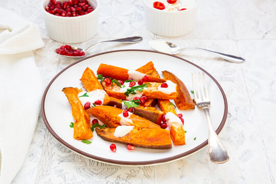 Plate of sweet potatoes with parsley, pomegranate seeds and cumin yogurt lying on white tiles