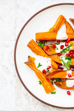 Plate of sweet potatoes with parsley, pomegranate seeds and cumin yogurt lying on white tiles