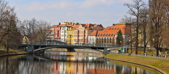 Park around Malse river in Ceske Budejovice with city center residential buildings in the background. The buildings reflect in the water.