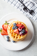 Homemade waffles with berries, powdered sugar and maple syrup on white plate, gray background.