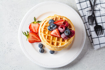 Homemade waffles with berries, powdered sugar and maple syrup on white plate, gray background.