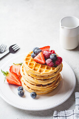 Homemade waffles with berries, powdered sugar and maple syrup on white plate, gray background.