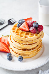 Homemade waffles with berries, powdered sugar and maple syrup on white plate, gray background.