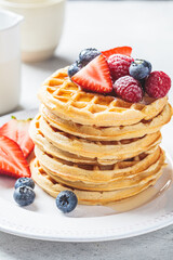Homemade waffles with berries, powdered sugar and maple syrup on white plate, gray background.