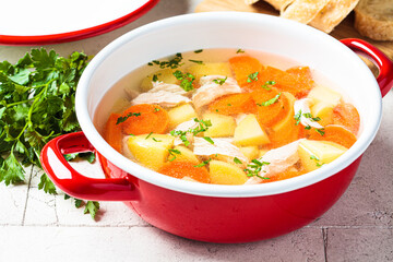 Traditional chicken soup with vegetables in red pot, gray background. Home cooking concept.