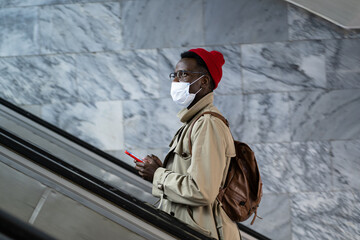 Serious African American young guy wear face mask for prevent coronavirus in public transport. Focused black hipster man in trench coat with mask as protection against covid-19 on escalator in subway
