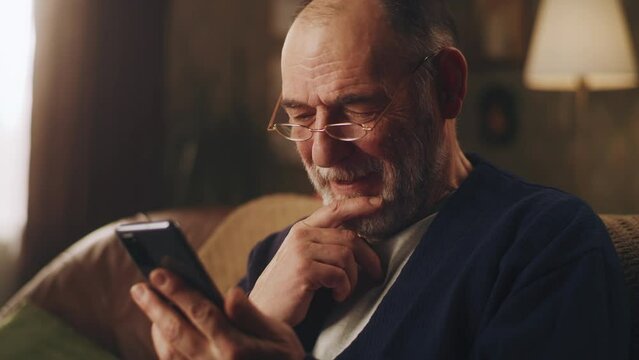 Elderly man smiling and watching funny videos on social networks using smartphone and resting in the evening sitting on the couch