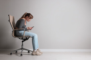 Young woman with poor posture using smartphone while sitting on chair near grey wall, space for text