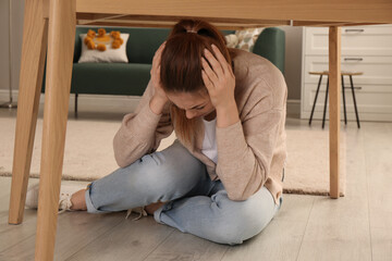 Scared woman hiding under table in living room during earthquake