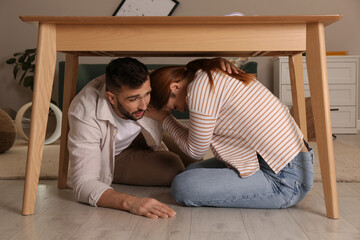 Scared couple hiding under table in living room during earthquake