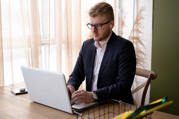 Blond caucasian young businessman wearing suit and glasses with headphones working with white laptop at desk in office or at home near big window. Man using computer