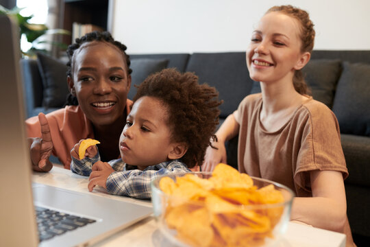 Joyful young mothers and son eating potato chips when watching movie on laptop screen