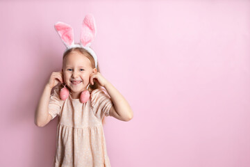 Obraz na płótnie Canvas Happy Easter. A cute beautiful girl with bunny ears holds green eggs in her hands as earrings. Pink background. Space for text