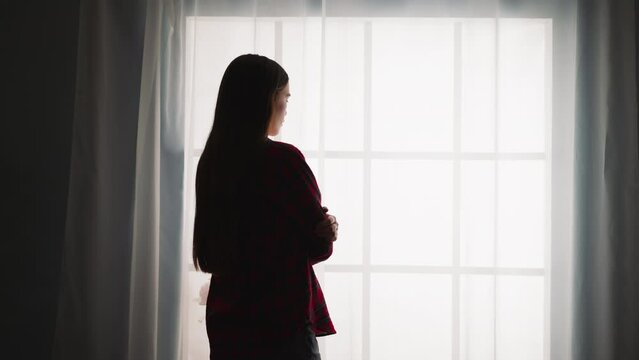 Silhouette of woman with long hair in shirt stands near French window in room backside view slow motion. Dreaming lady spends time alone at home