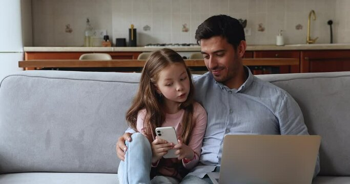 Cute preschool 6s girl holds smartphone seated on sofa with young loving dad with notebook on laps, family use diverse devices spend weekend on internet. Young generation gadgets overuse, apps concept