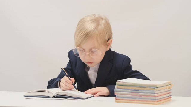 Boy of 7-8 years old in large glasses wears jacket and shirt sits at desk and writes in notebook. Child behaves like boss or director.