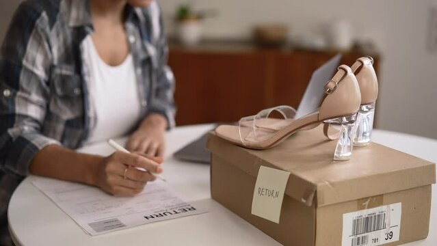 Young woman writing return order and sitting at table with shoes in home room spbd. 4k Closeup view of american african female customer writes on paper and wants to return goods back, sits at desk