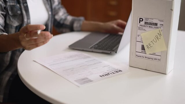 Young woman using laptop and writing refund from order at table in home spbd. 4k Close view of female client types on computer and writes on paper document, wants to return goods and sits at desk with
