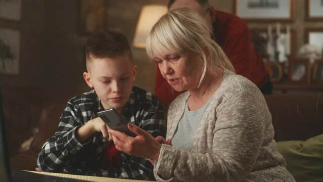 A teen grandson teaching his aged grandparents how to use the application on a modern smartphone while sitting on the sofa in a family house