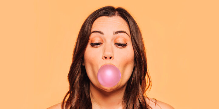 Young beautiful girl inflates a pink chewing gum. Youngster female in the studio on a beige background. The woman's face with bright black shadow on the eyes. Copy space.