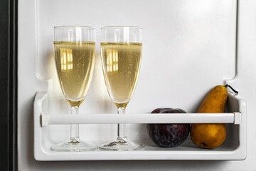 Two clean shiny glasses of champagne or sparkling wine with window reflection with natural ripe fruits: plum and pear on white fridge opened door in daylight close up. Celebration, life event, holiday