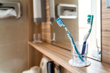 Two used blue and turquoise toothbrushes in glass cup stand on wooden shelf near mirror and window...