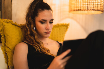 Chica joven delgada con pelo largo mirando tablet sobre la cama de un hotel estilo soho