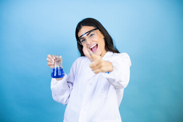 Young brunette woman wearing scientist uniform holding test tube over isolated blue background pointing to you and the camera with fingers, smiling positive and cheerful