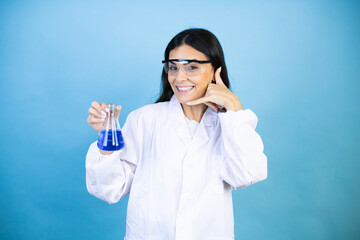 Young brunette woman wearing scientist uniform holding test tube over isolated blue background smiling doing phone gesture with hand and fingers like talking on the telephone