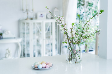 Blooming tree branches in the vase and colored easter eggs on the plate on white kitchen table with classic style interior background. Easter celebration at home. Festive spring composition.