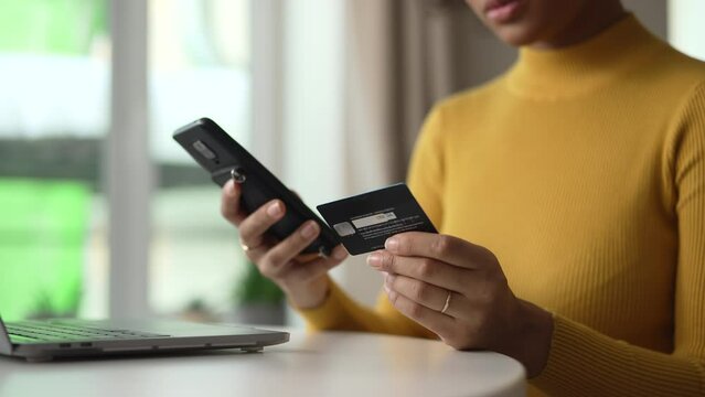 Young woman using devices and credit card for online shopping and sitting at table at home spbd. 4k Closeup of american african female holds smartphone and looks with smile, enters payment data and