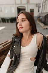 Fototapeta na wymiar Fashionable beautiful stylish woman model hipster with short hair in a fashion black leather jacket with a white dress sits and relaxes in the city on wooden pallets