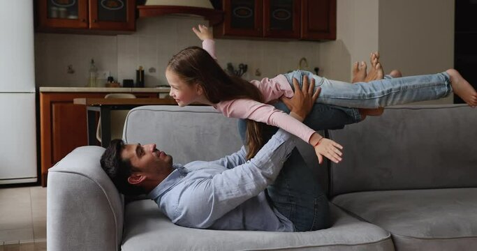 Loving stepdad lying on sofa play with little daughter, preschool girl spread arms pretend flying in air enjoy playtime with caring young dad. Father Day, happy fatherhood, family game at home concept