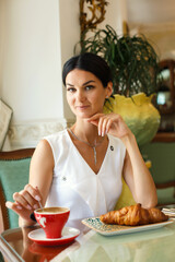 A beautiful young woman with black hair poses in a white dress in a luxury hotel or restaurant. The girl is sitting at a cafe table and holding a cup of coffee in her hand